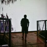 A silhouette of a person walking towards a large abstract light display indoors, creating a dramatic contrast.